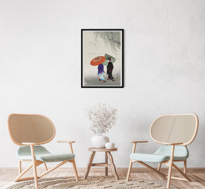 A framed poster of 'Two Women in the Rain' by Ohara Koson, displayed on a wall above a small round table with a vase, next to two modern chairs.